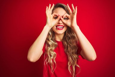 Genç ve güzel bir kadın, kırmızı izole edilmiş arka plan üzerinde duran tişört giyiyor. Dürbün gibi güzel hareketler yapıyor. Gözleri parmaklarından dışarı bakıyor. Çılgın ifade.