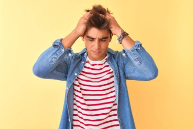 Çizgili tişört ve kot gömlek giyen genç yakışıklı adam izole edilmiş sarı arka planda acı ve migren yüzünden çaresiz ve stresli bir baş ağrısından muzdarip. Ellerini başının üstüne koy..