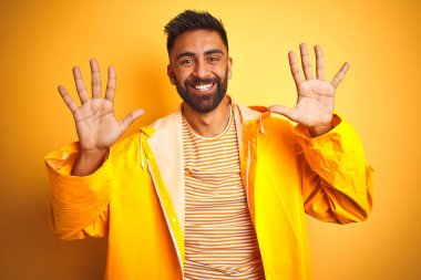 İzole edilmiş sarı arka planda duran yağmurluk giyen genç Hintli adam on numaralı parmaklarıyla işaret ederken kendinden emin ve mutlu gülümsüyor..