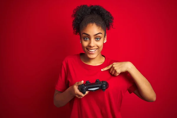 非洲裔美国女游戏玩家用操纵杆在孤立的红色背景上玩电子游戏 惊讶地将手指指向自己 — 图库照片