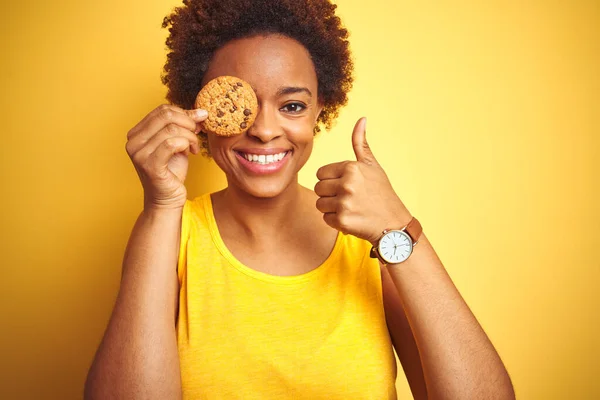 African american woman holding chocolate chips cookie over yellow background happy with big smile doing ok sign, thumb up with fingers, excellent sign