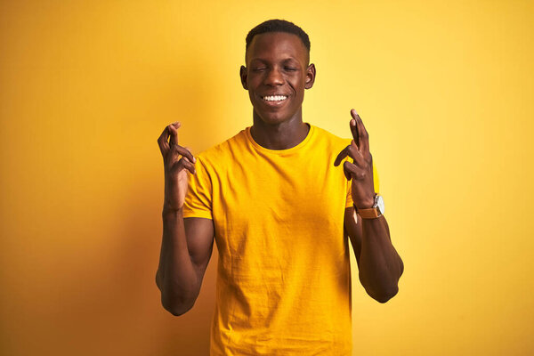 Young african american man wearing casual t-shirt standing over isolated yellow background gesturing finger crossed smiling with hope and eyes closed. Luck and superstitious concept.