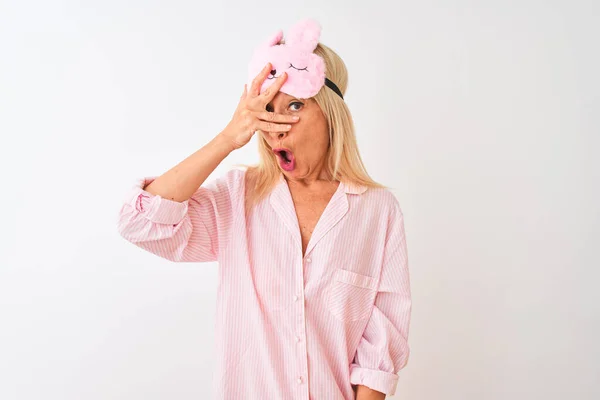 Middle age woman wearing sleep mask and pajama over isolated white background peeking in shock covering face and eyes with hand, looking through fingers with embarrassed expression.