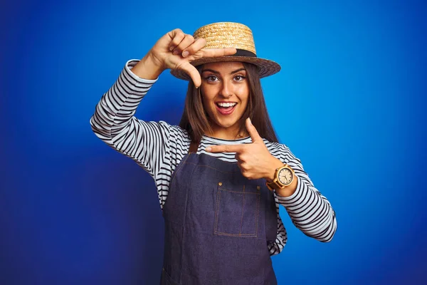 Young beautiful baker woman wearing apron and hat standing over isolated blue background smiling making frame with hands and fingers with happy face. Creativity and photography concept.