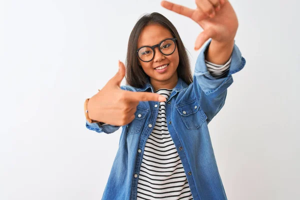 Young chinese woman wearing denim shirt and glasses over isolated white background smiling making frame with hands and fingers with happy face. Creativity and photography concept.