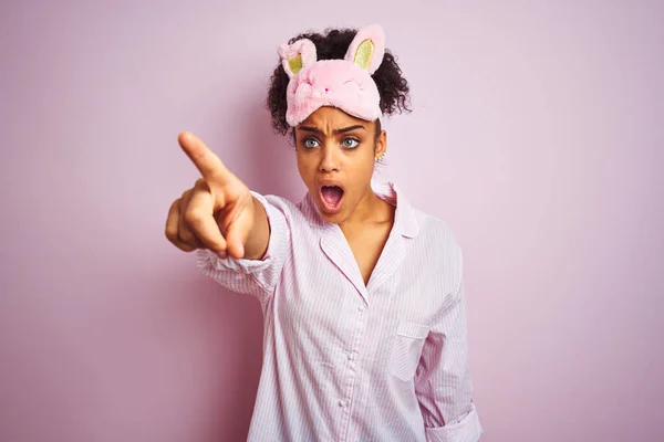 年轻的非洲裔美国女人 戴着睡衣和面罩 罩在孤立的粉色背景上 手指尖尖着 前面是惊讶的样子 嘴角是惊讶的表情 前面是什么东西 — 图库照片