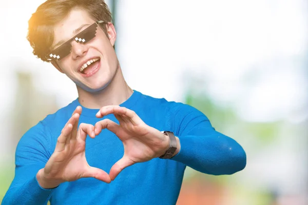 Young man wearing funny thug life glasses over isolated background smiling in love showing heart symbol and shape with hands. Romantic concept.
