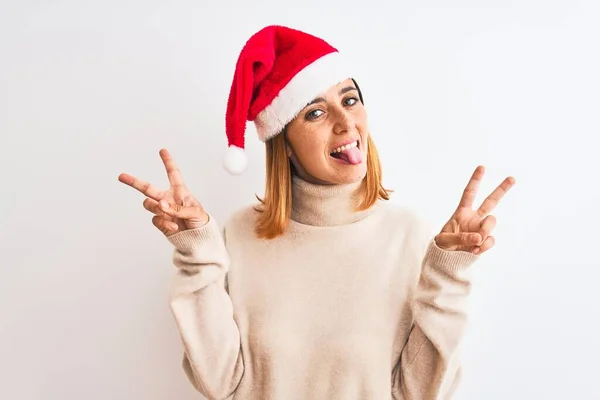 孤立した背景の上にクリスマスの帽子を身に着けている美しい赤毛の女性は勝利の印をする両方の手の指を示す舌で笑っている 第二番 — ストック写真