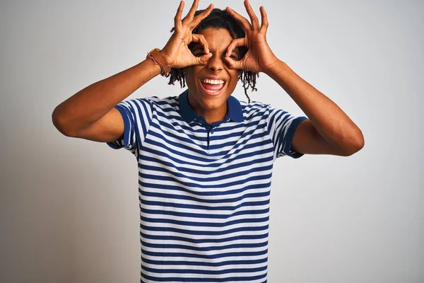 Afro man with dreadlocks wearing striped blue polo standing over isolated white background doing ok gesture like binoculars sticking tongue out, eyes looking through fingers. Crazy expression.