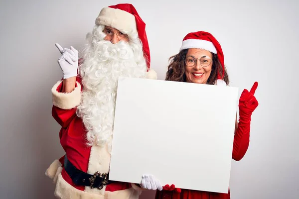 上了年纪的夫妇穿着圣诞老人的服装 拿着横幅在孤立的白色背景上 很高兴手指手画脚地指向旁边 — 图库照片