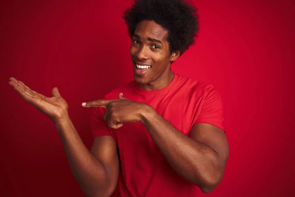 Young american man with afro hair wearing t-shirt standing over isolated red background amazed and smiling to the camera while presenting with hand and pointing with finger.