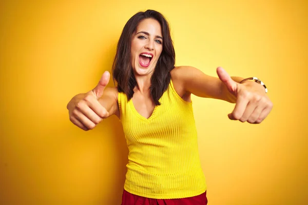 Young beautiful woman wearing t-shirt standing over yellow isolated background approving doing positive gesture with hand, thumbs up smiling and happy for success. Winner gesture.