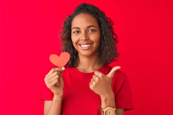 年轻的巴西女人拿着纸心站在孤立的红色背景上 开心的笑着做着好的手势 用手指竖起大拇指 很好的标志 — 图库照片
