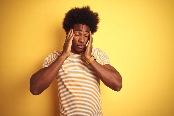 American man with afro hair wearing striped t-shirt standing over isolated yellow background with hand on headache because stress. Suffering migraine.