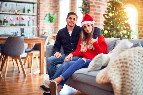 一对年轻的夫妇头戴圣爪帽 坐在沙发上围着圣诞树 脸上带着严肃的表情 简单而自然地看着相机 — 图库照片