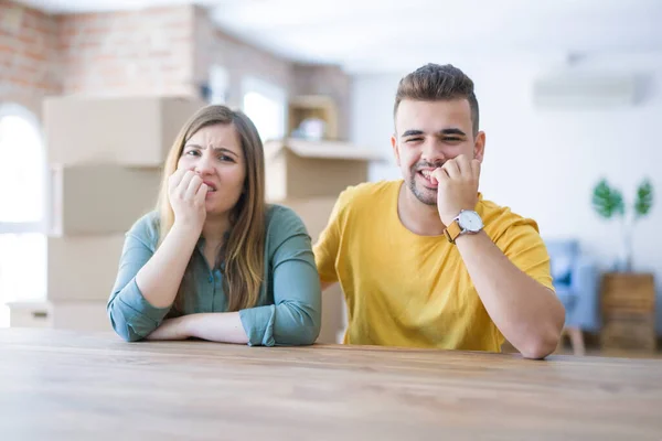 Young couple sitting on the table movinto to new home with carboard boxes behind them looking stressed and nervous with hands on mouth biting nails. Anxiety problem.