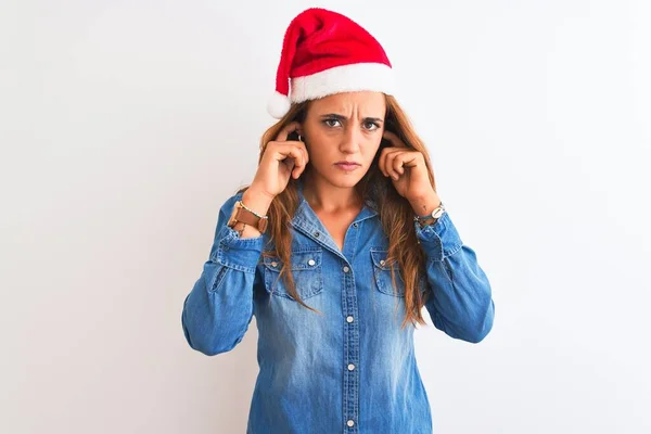 孤立した背景の上にクリスマスの帽子をかぶっている若い美しい赤毛の女性は 大きな音楽の騒音のためにいらいらする表情で耳を覆います ろう者の概念 — ストック写真