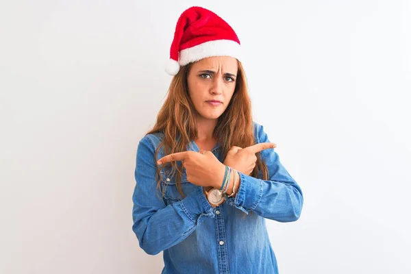 若いです美しい赤毛女性は孤立した背景の上にクリスマスの帽子を身に着けています指で両側に指しています 異なる方向は反対します — ストック写真