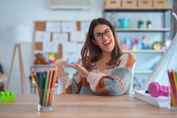 Young beautiful teacher woman wearing sweater and glasses sitting on desk at kindergarten Inviting to enter smiling natural with open hand
