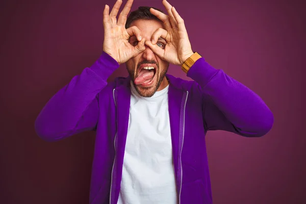 Young fitness man wearing casual sports sweatshirt over purple isolated background doing ok gesture like binoculars sticking tongue out, eyes looking through fingers. Crazy expression.