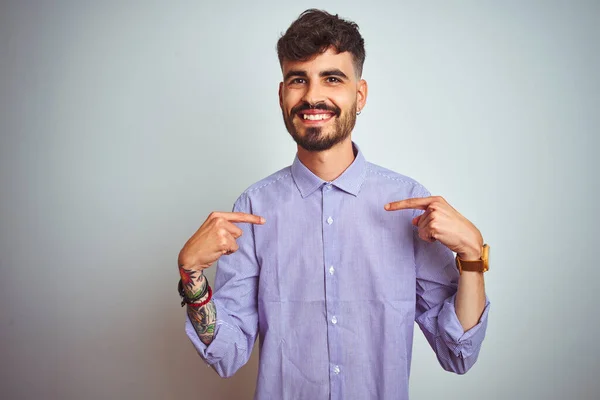 Young man with tattoo wearing purple shirt standing over isolated white background looking confident with smile on face, pointing oneself with fingers proud and happy.