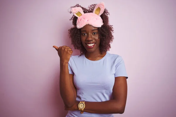 Young african afro woman wearing sleep mask standing over isolated pink background smiling with happy face looking and pointing to the side with thumb up.