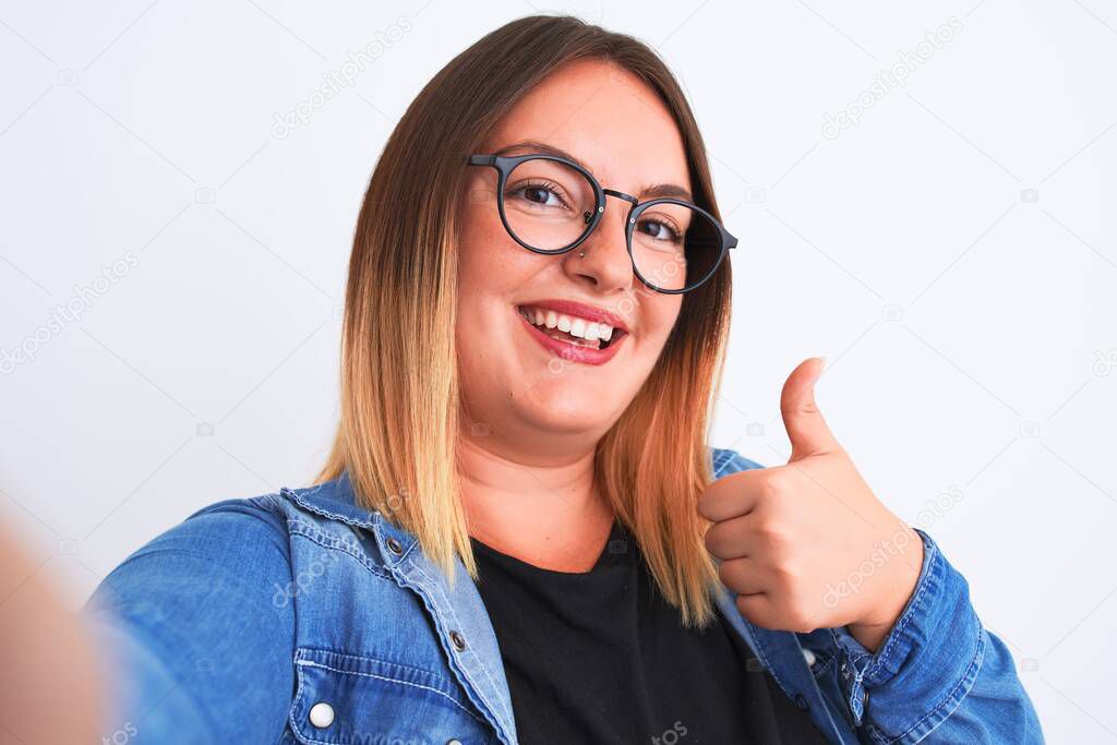 Beautiful woman wearing denim shirt and glasses make selfie over isolated white background happy with big smile doing ok sign, thumb up with fingers, excellent sign