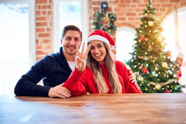 Noel Baba şapkası takan genç bir çift Noel ağacının etrafında oturmuş gülümseyen ve mutlu bir şekilde üç numaralı parmaklarıyla işaret ediyor..