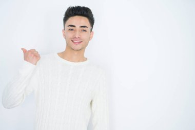 Yakışıklı genç bir adam, izole edilmiş beyaz bir arka planda gülümsüyor, gülümsüyor ve başparmağıyla yana bakıyor..