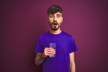 Dövmeli genç adam, izole edilmiş mor arka planda duran bir bardak su içiyor. Şaşırmış bir yüzle korkmuş ve korku ifadesinden heyecanlanmış.