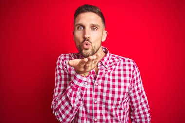 Kırmızı izole edilmiş arka plan üzerinde genç yakışıklı bir adam kameraya bakıp havada bir öpücük savuruyor ve sevimli ve seksi oluyor. Aşk ifadesi.