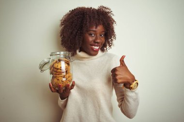 Afrikalı genç bir afro kadın, elinde bir kavanoz kurabiyeyle izole edilmiş beyaz arka planda mutlu bir şekilde duruyor. Gülümsüyor, işaret veriyor, parmaklarıyla başparmağını kaldırıyor.