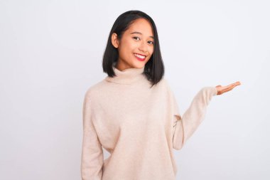 Balıkçı yaka kazak giyen genç bir kadın izole edilmiş beyaz arka planda dikiliyor neşeli bir şekilde gülümsüyor ve avuç içi kameraya bakıyor..