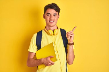 Kulaklık takan ve sırt çantası olan genç bir çocuk izole bir arka planda kitap okuyor. Çok mutlu bir şekilde el ve parmak uçlarını işaret ediyor.