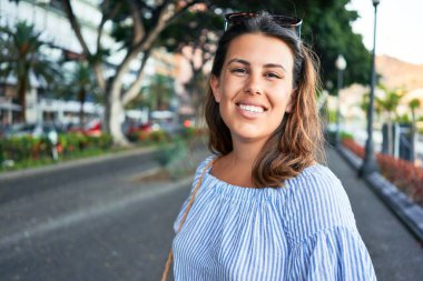 Genç ve güzel bir kadın mutlu bir şekilde gülümsüyor güneşli bir yaz gününde şehir sokaklarında yürüyor.