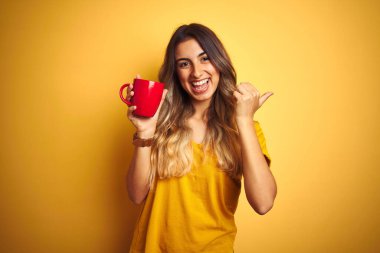 Sarı arka planda kırmızı kahve bardağını tutan genç ve güzel bir kadın başparmağıyla gülümseyerek yüzünü gösteriyor.