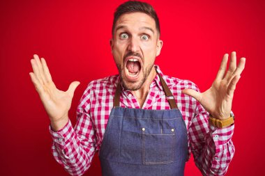 Kırmızı izole edilmiş arka plan üzerinde dükkan sahibi önlüğü giyen genç yakışıklı adam çılgın ve başarıyı kollarını kaldırıp heyecanla haykırarak kutluyor. Kazanan konsept