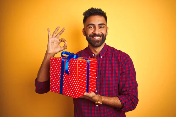 Arab indian hispanic man holding birthday gift standing over isolated yellow background doing ok sign with fingers, excellent symbol