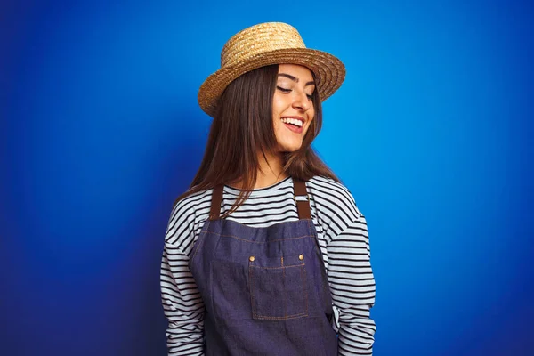 Young beautiful baker woman wearing apron and hat standing over isolated blue background looking away to side with smile on face, natural expression. Laughing confident.