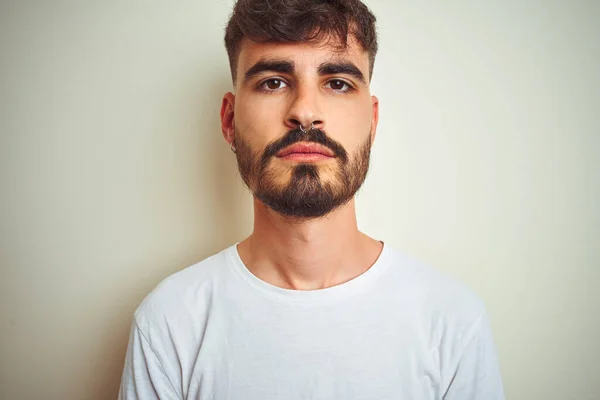 Young man with tattoo wearing t-shirt standing over isolated white background with serious expression on face. Simple and natural looking at the camera.