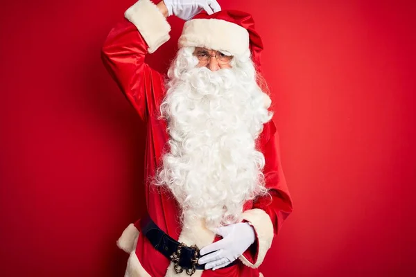 Middle age handsome man wearing Santa costume standing over isolated red background confuse and wondering about question. Uncertain with doubt, thinking with hand on head. Pensive concept.