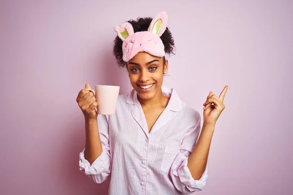 アフロ女性身に着けていますPajamaとマスク飲みますコーヒーカップ上の隔離されたピンクの背景非常に幸せなポインティングで手と指で側面 — ストック写真