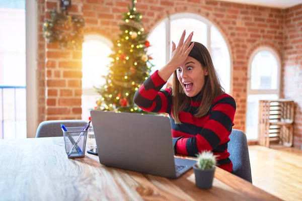 Beautiful woman sitting at the table working with laptop at home around christmas tree surprised with hand on head for mistake, remember error. Forgot, bad memory concept.