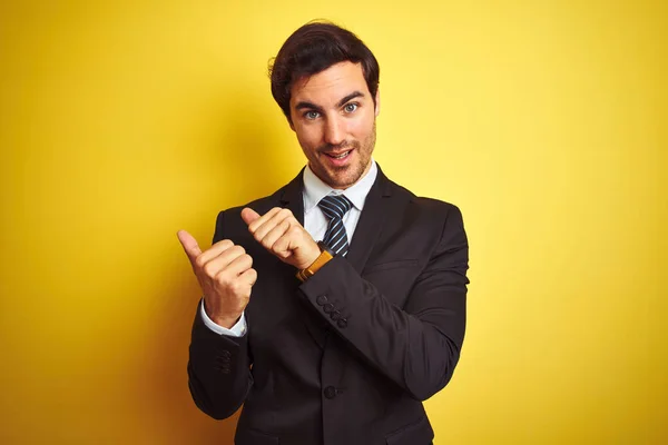 若いハンサムなビジネスマンがスーツを着て 孤立した黄色の背景の上に立ってネクタイ手と親指を後ろに向け 自信を持って笑って — ストック写真