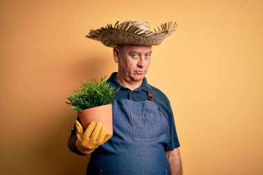 Orta yaşlı, beyaz önlüklü ve şapkalı bir çiftçi sarı arka planda bitki saksısı tutuyor. Zeki bir yüz ifadesiyle ciddi ciddi düşünüyor.