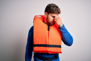 Sakallı, mavi gözlü, beyaz arka planda can yeleği giyen, burnu ve gözleri ovuşturan yorgun, bitkin ve baş ağrısı çeken sarışın genç bir turist. Stres ve hüsran kavramı.