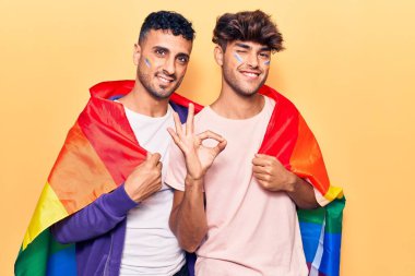 Gökkuşağı rengi lgbtq bayrağı giyen genç eşcinsel çift parmaklarıyla işaret veriyor, gülümseyerek mükemmel bir sembol oluşturuyor. 
