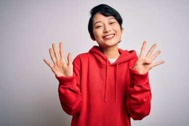 Genç, güzel Asyalı bir kız beyaz arka planda kapüşonlu kazak giyer ve dokuz numarayı göstererek gülümserken kendinden emin ve mutlu görünür..