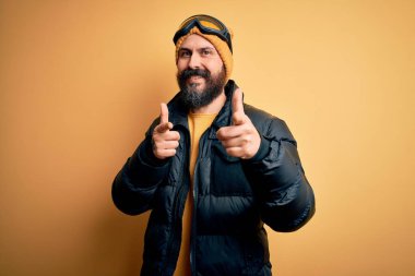 Sakallı, kar maskesi takmış, kar maskesi takmış ve kayak gözlüklü yakışıklı kayakçı mutlu ve komik yüzüyle kamerayı işaret ediyor. İyi enerji ve titreşimler.