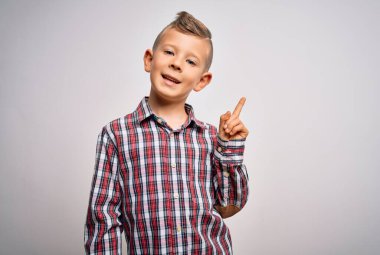 Mavi gözlü, zarif gömlekli küçük beyaz çocuk izole edilmiş arka planda duruyor. Yüzünde kocaman bir gülümseme var. Kameraya el parmağı ile bakıyor..